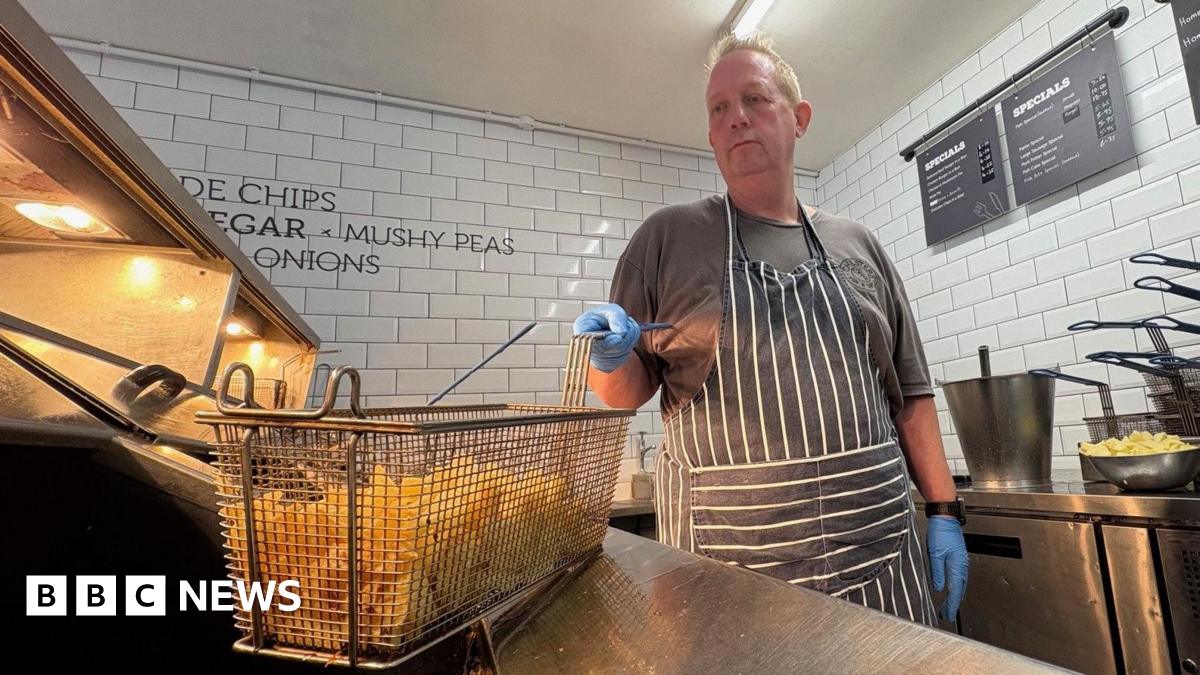 Paul Costelloe has receding grey hair. He is wearing a bright pink, blue and green scarf and a blue jacket.