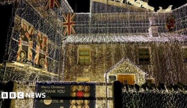 Richard Iles' house completely covered in white string lights. A sign attached to a gate says Merry Christmas and a thank you message to veterans. Union Jack flags are also hung from his home.
