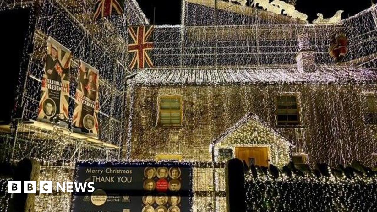 Richard Iles' house completely covered in white string lights. A sign attached to a gate says Merry Christmas and a thank you message to veterans. Union Jack flags are also hung from his home.