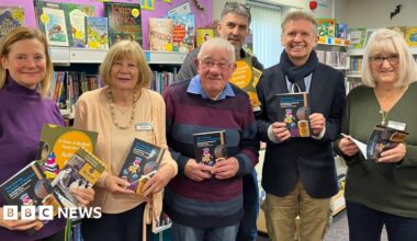 Six people in a line. They hold up copies of the same book and are all smiling at the camera. They are in a library with a shelf full of colourful books is behind them.