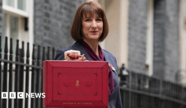 Rachel Reeves, a woman with dark brown hair, dressed in a grey suit with purple scarf, holds up a red box with gold coloured handle and gold coloured lettering in street with black railings behind her.