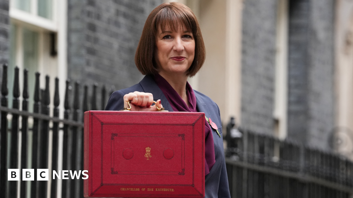 Rachel Reeves, a woman with dark brown hair, dressed in a grey suit with purple scarf, holds up a red box with gold coloured handle and gold coloured lettering in street with black railings behind her.