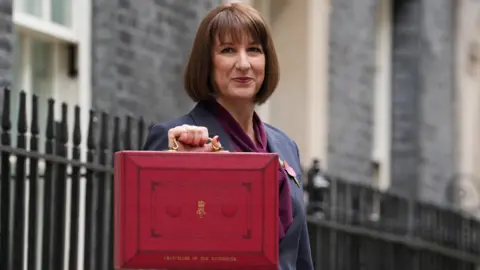 Reuters Rachel Reeves, a woman with dark brown hair, dressed in a grey suit with purple scarf, holds up a red box with gold coloured handle and gold coloured lettering in street with black railings behind her.