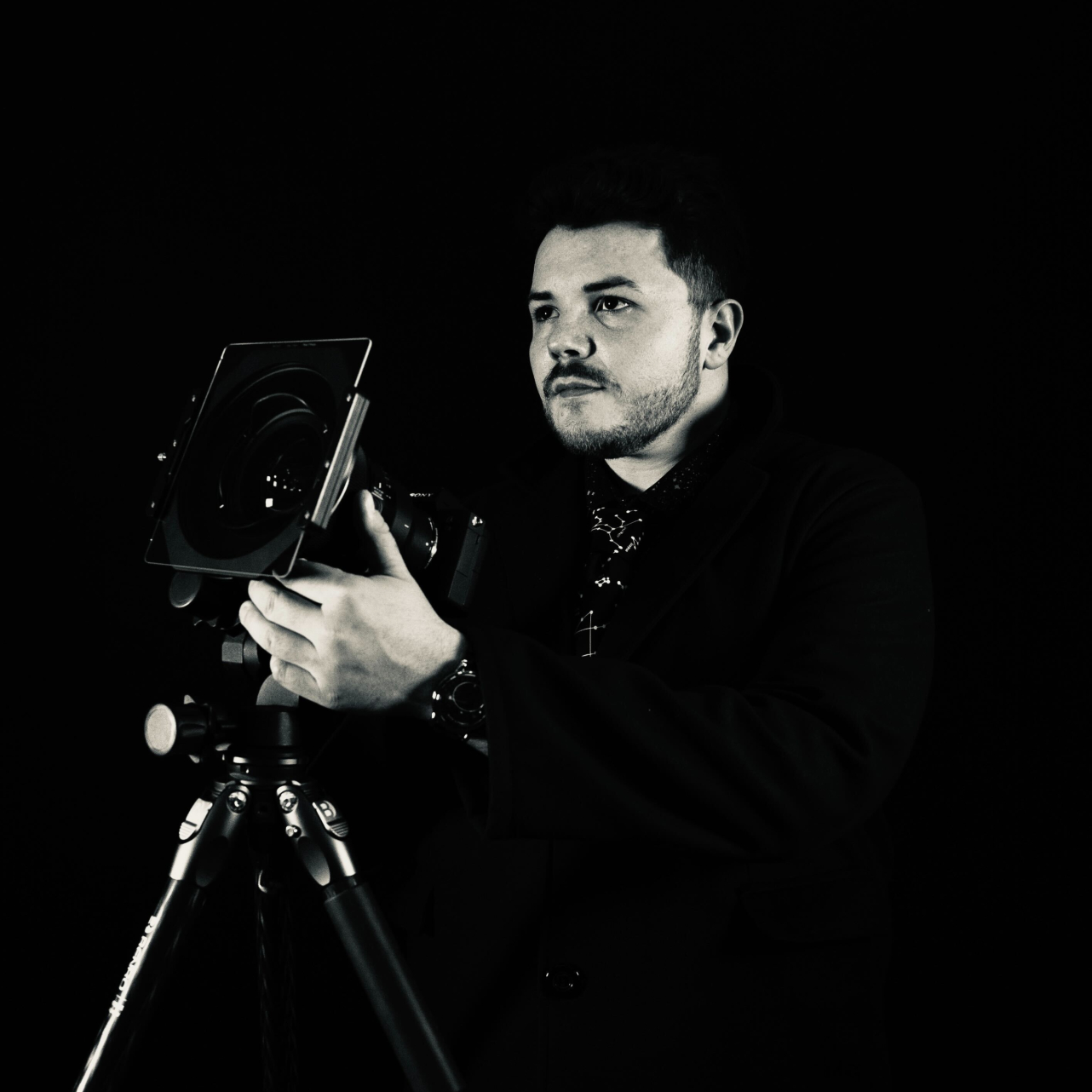 A man wearing all black holds a camera on a tripod in a black and white photo