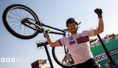 Charlie Rolls smiles at the camera as he holds one of his arms up in the air in celebration. In his other hand he holds his bike up in the air. He wears a black helmet with a white top that has GBR written on the chest and he wears black shorts as well as gloves. He has some stubble on his face.