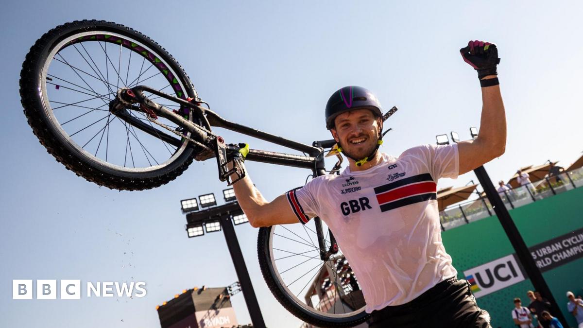 Charlie Rolls smiles at the camera as he holds one of his arms up in the air in celebration. In his other hand he holds his bike up in the air. He wears a black helmet with a white top that has GBR written on the chest and he wears black shorts as well as gloves. He has some stubble on his face.