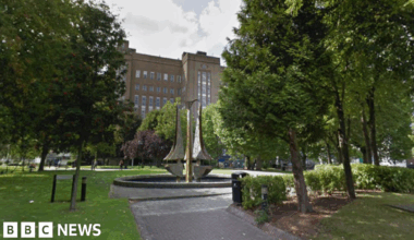 An early 20th-Century brick building, approximately six stories high, is flanked by trees and green space with a modern sculpture, Tipping Triangles, in a water feature in the foreground. The sculpture consists of three 18-foot stainless steel "triangles" with one curved side.