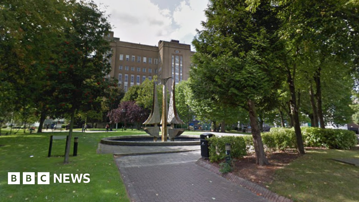 An early 20th-Century brick building, approximately six stories high, is flanked by trees and green space with a modern sculpture, Tipping Triangles, in a water feature in the foreground. The sculpture consists of three 18-foot stainless steel "triangles" with one curved side.