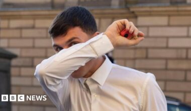 Liam McDermid covering his face with his right arm. He is holding a red and yellow vape in his right hand. He has dark hair and is wearing a white shirt.