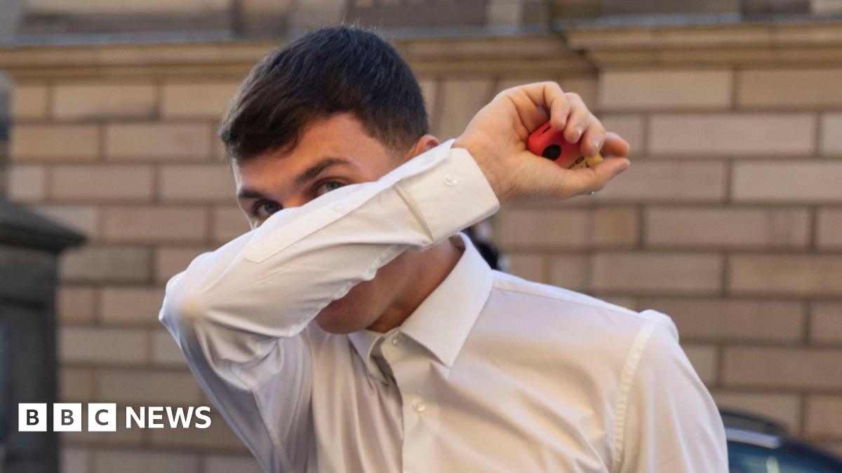 Liam McDermid covering his face with his right arm. He is holding a red and yellow vape in his right hand. He has dark hair and is wearing a white shirt.