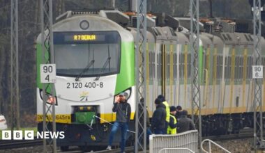 A green-and-white train stopped on a railway track, with officials standing beside it. Overhead power lines and trackside signs are visible in the scene.