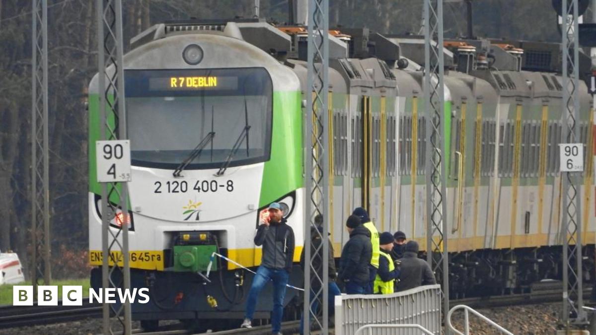 A green-and-white train stopped on a railway track, with officials standing beside it. Overhead power lines and trackside signs are visible in the scene.