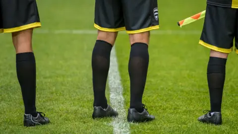 Getty Images The lower bodies of three football match officials standing on grass. The photo is taken from behind and they are wearing black and yellow kit.