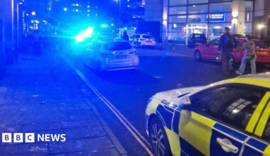 A blurry image of a city centre street with several police cars parked up with their blue lights on. There is a police station in the background and a number of people stood outside.