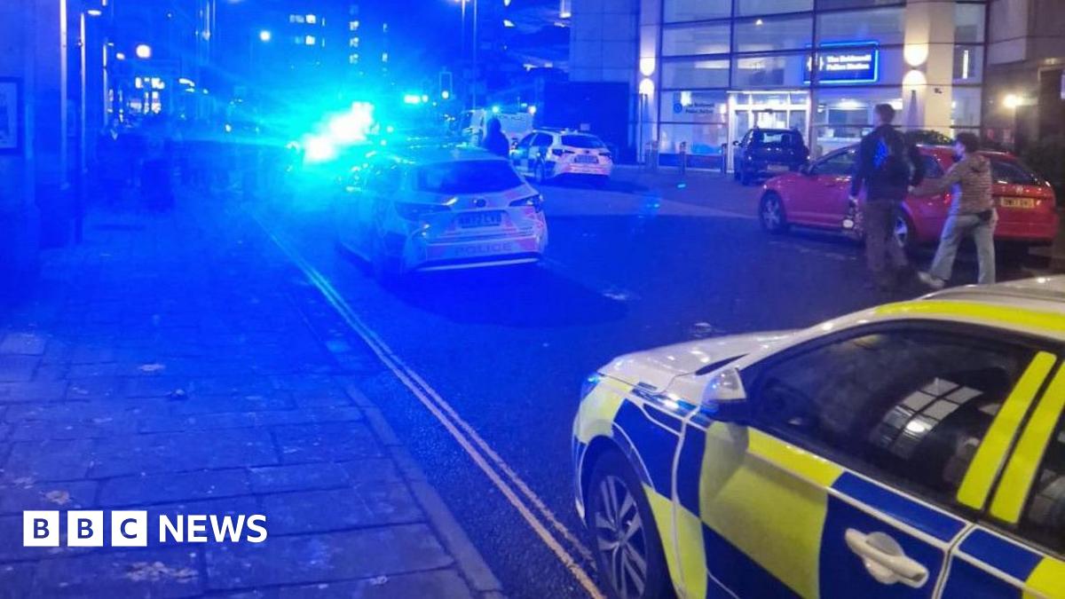 A blurry image of a city centre street with several police cars parked up with their blue lights on. There is a police station in the background and a number of people stood outside.