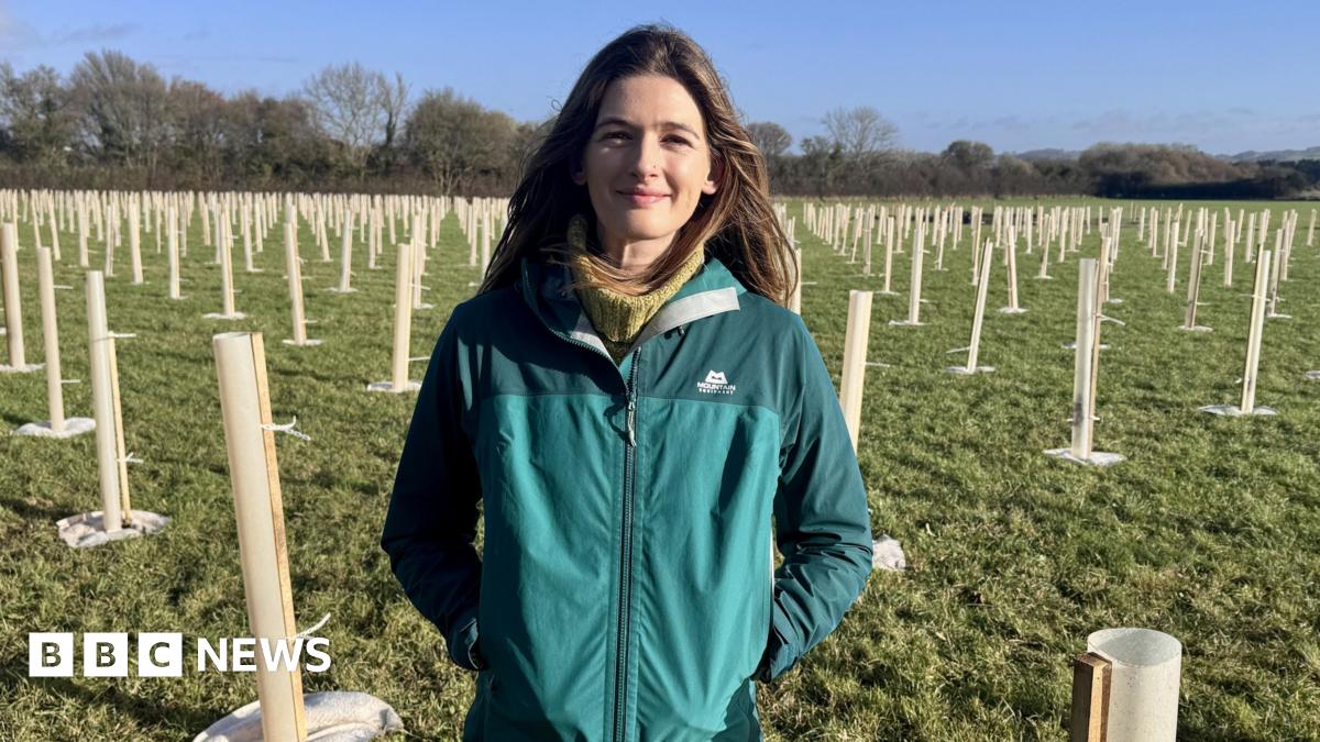A woman with long brown hair standing in the middle of a woodland where hundreds of trees are being grown. She is wearing a blue outdoors jacket with her hands in her pockets.