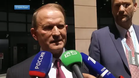 Channel One Russia A man with brown hair wearing a blue pinstripe suit and claret tie talks into three microphones of Russian television - two blue and one green - while conducting a press briefing outside the European Parliament building. A tall man with a blue suit and red tie is standing next to him 