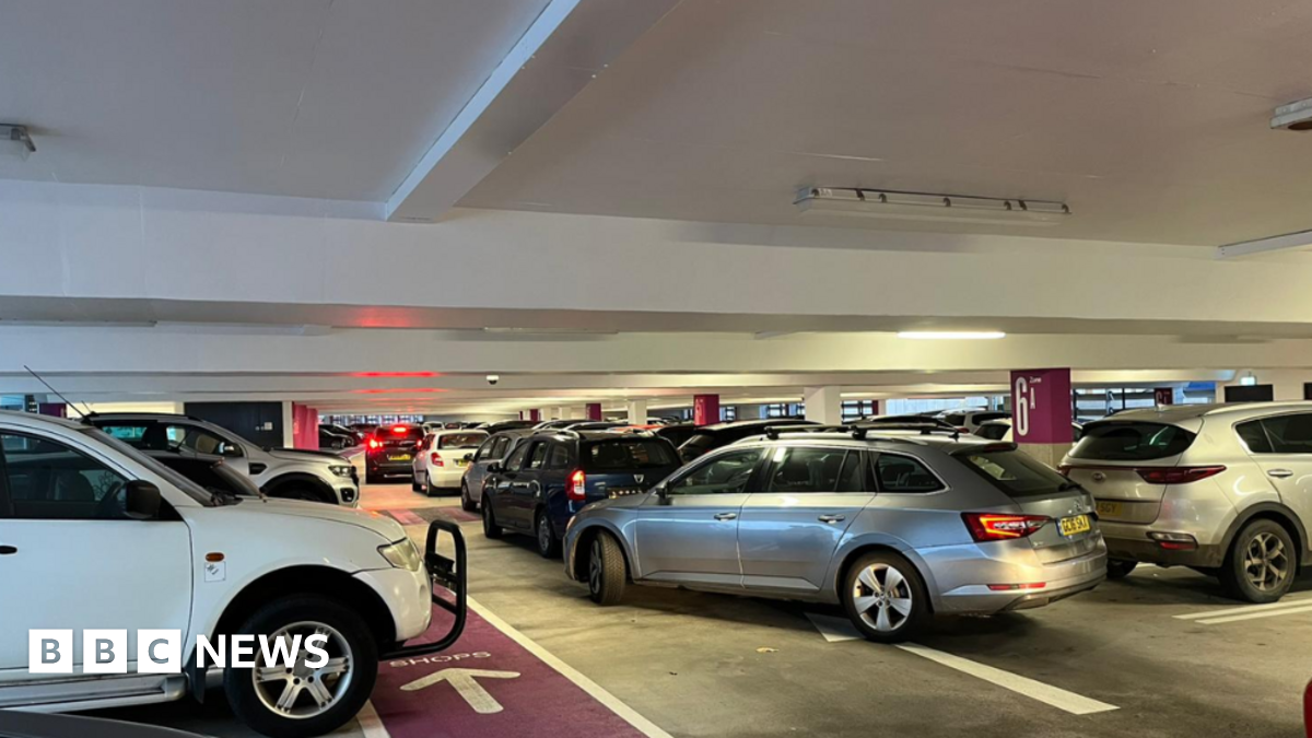 A line of cars trying to leave a high-rise car park. The ceiling is white and there are white lines and road markings painted on the floor.