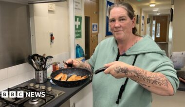 A woman is stood in a kitchen at a community centre. Her hair is tied back and she is wearing a green hoodie. She is holding up a frying pan full of sausages