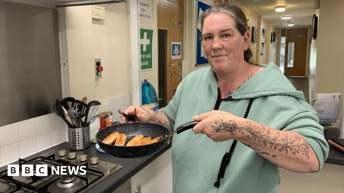 A woman is stood in a kitchen at a community centre. Her hair is tied back and she is wearing a green hoodie. She is holding up a frying pan full of sausages