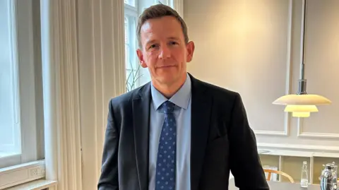 Rasmus Stoklund a middle aged man with short blonde hair wearing a suit and tie, next to a window in his office