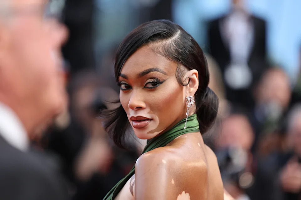 Person on the red carpet in elegant, form-fitting attire, looking over their shoulder with glamorous earrings and sleek hairstyle
