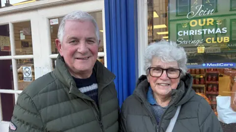 Mr and Mrs Linton are standing beside each other in a street. They're both wearing grey jackets. Mrs Linton wears glasses. A business advertising their Christmas club is behind them. 
