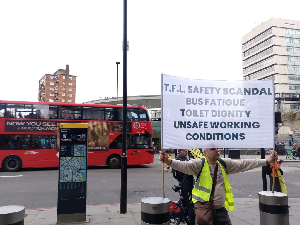 A London bus driver holds a banner that reads "TfL safety scandal, bus fatigue, toilet dignity, unsafe working conditions' as a red double-decker bus passes in the background. 