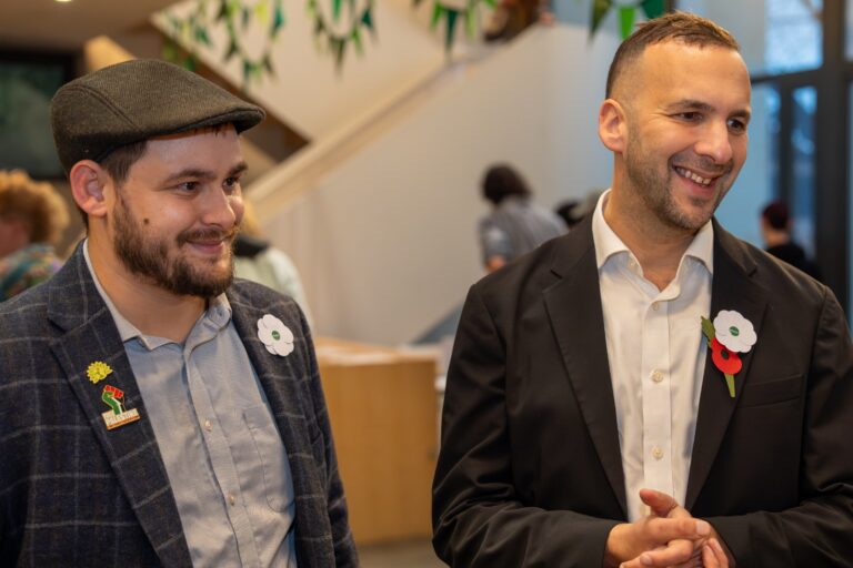 Two men in suits stand next to each other smiling.