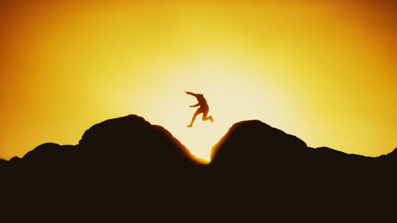 A person jumps between two rocky cliffs at sunset, silhouetted against a bright, golden sky.
