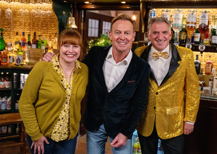 Jason Donovan stands between Emmerdale's Lydia Dingle and Bob Hope - who is wearing a glittery, golden suit - on The Woolpack set in Emmerdale