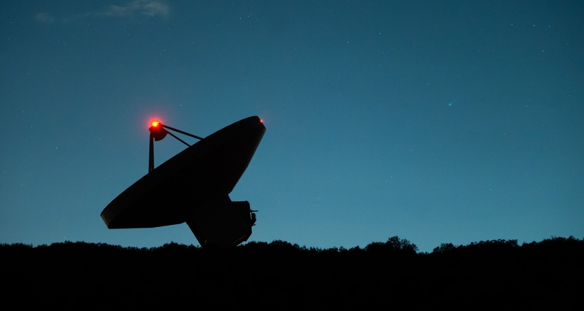 A dark silhouette of a satellite dish tilts to the left next to the dark horizon with a dark blue night sky in the background