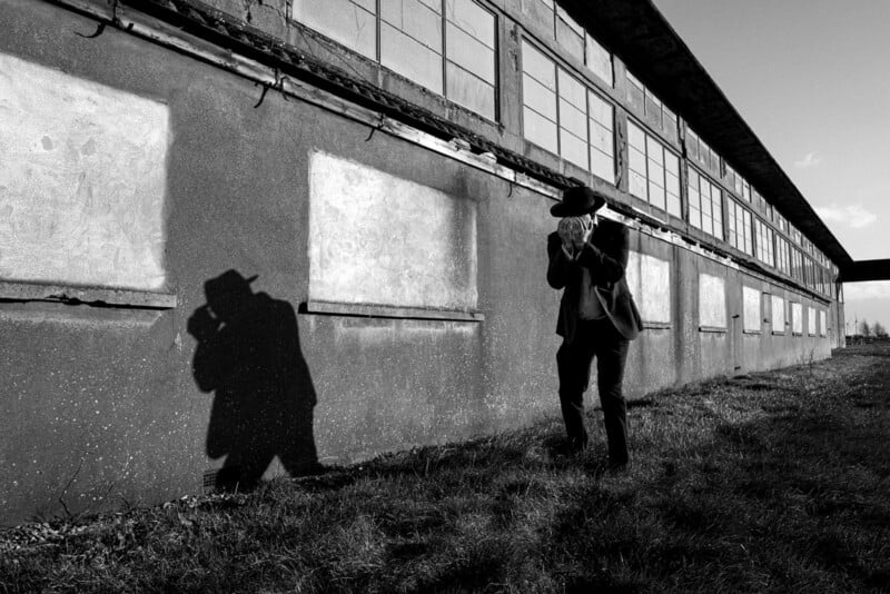 A person in a hat walks along the exterior wall of an industrial building, casting a distinct shadow on the wall. The scene is in black and white, with sunlight creating strong contrasts and long shadows on the grass.