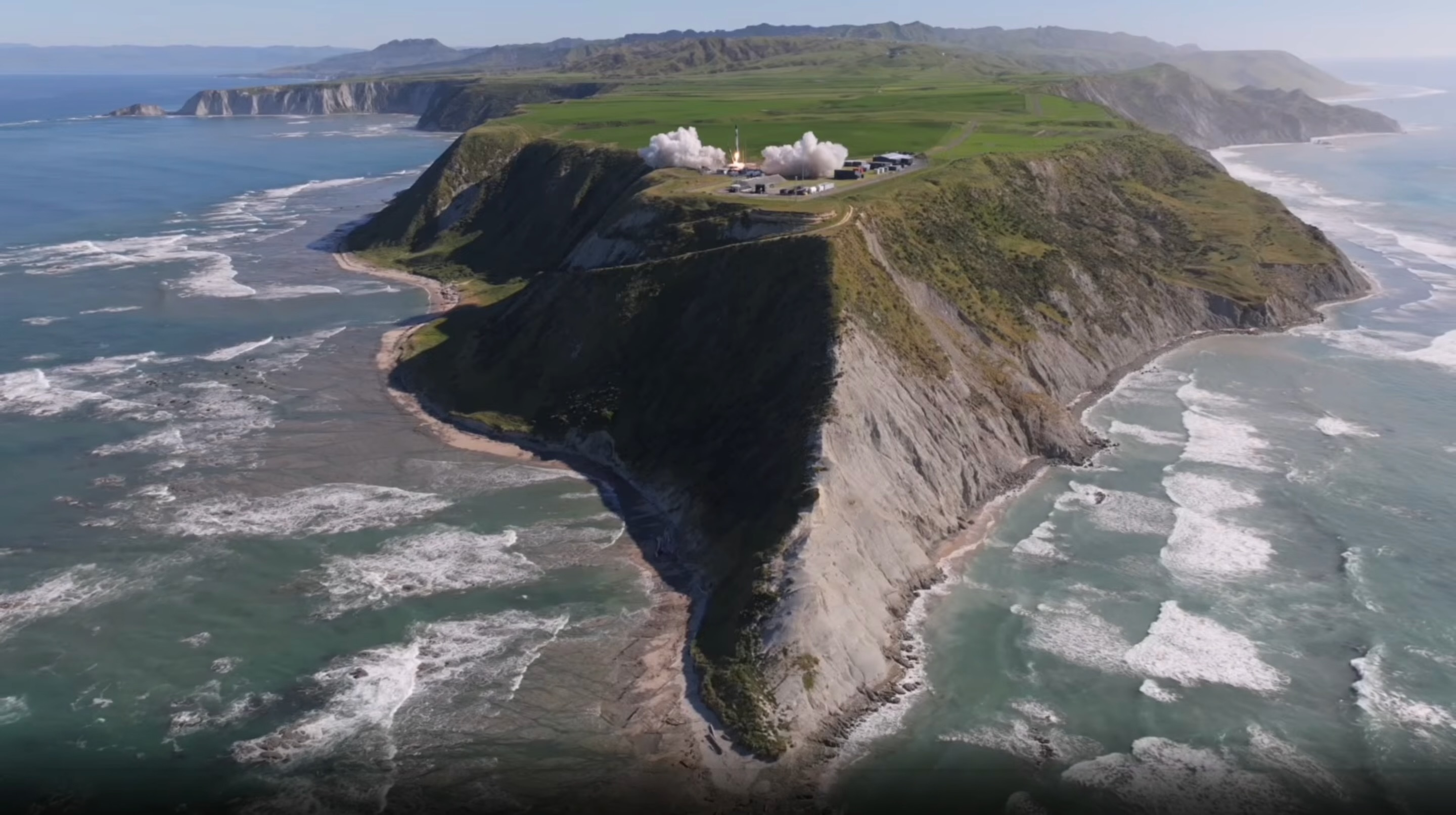 a plume of grey smoke forms on the tip of a rocky island rising out of a wavy ocean