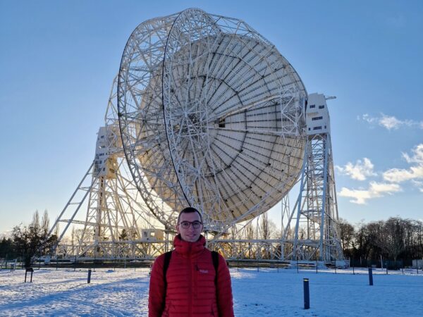 A young man in a red jacket standing in the snow with a radio dish behind him.