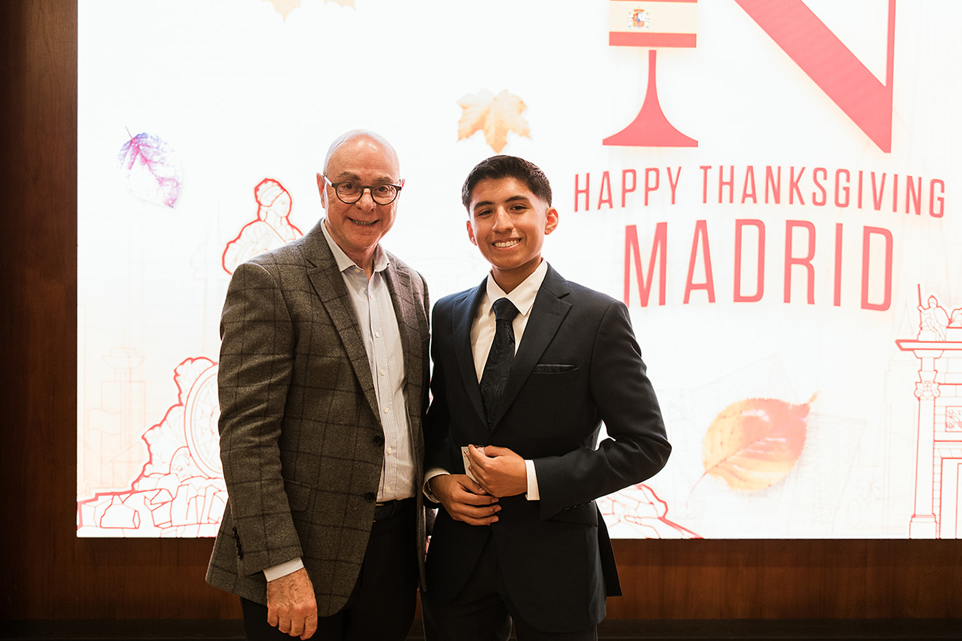 Northeastern University president Joseph Aoun with a suited man in front of a sign saying 'Happy Thanksgiving Madrid"