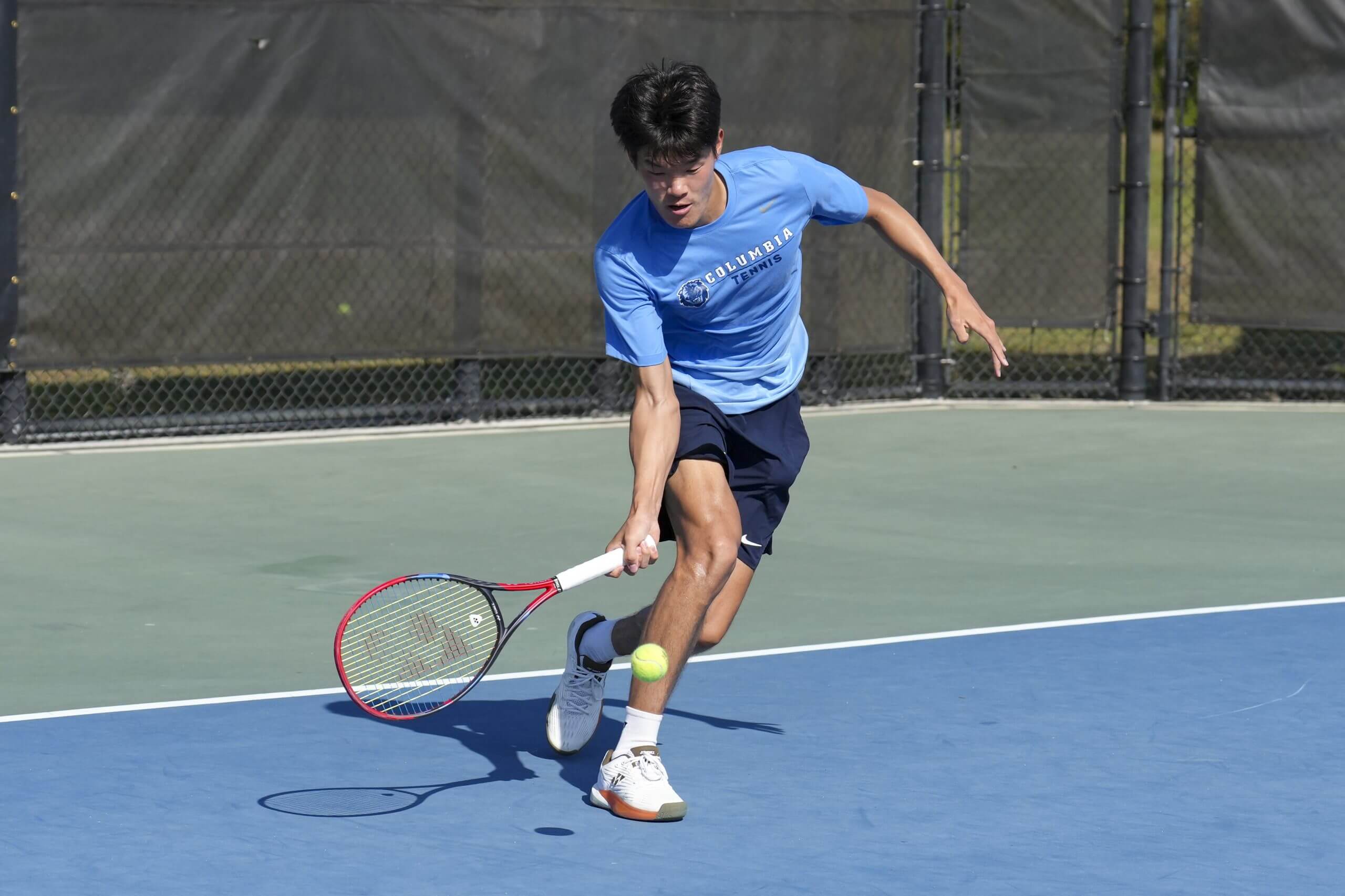 Michael Zheng bends low to hit a forehand slice on a blue tennis court, wearing a Columbia T shirt.