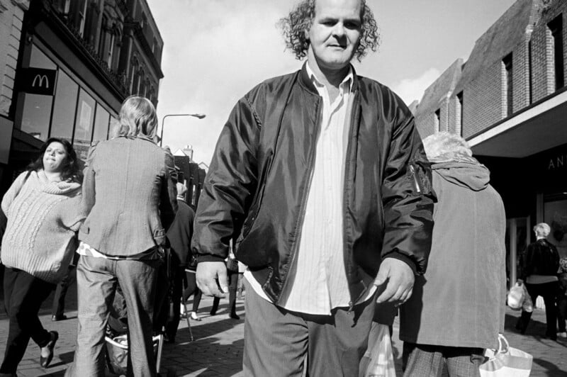 A man with curly hair and a jacket stands in the foreground on a busy city street, surrounded by people walking in various directions. There are shops and buildings in the background and a partly cloudy sky above.