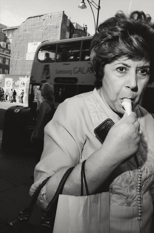 A woman holding a phone and bag eats an ice cream on a city street. A double-decker bus and pedestrians are visible in the background on a sunny day.