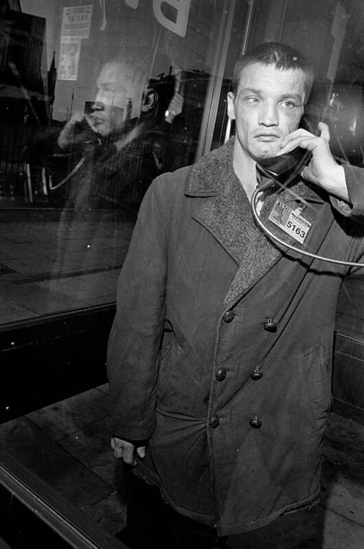 A man in a heavy coat stands in a phone booth holding a payphone receiver to his ear. His reflection is visible in the glass behind him. The expression on his face appears somber or contemplative.