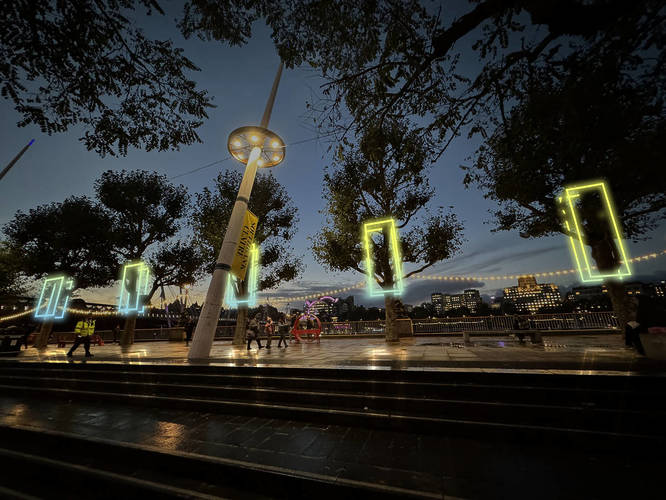 Geometric shapes of bright colourful light frame the trunks of the trees that line the riverside walkway.