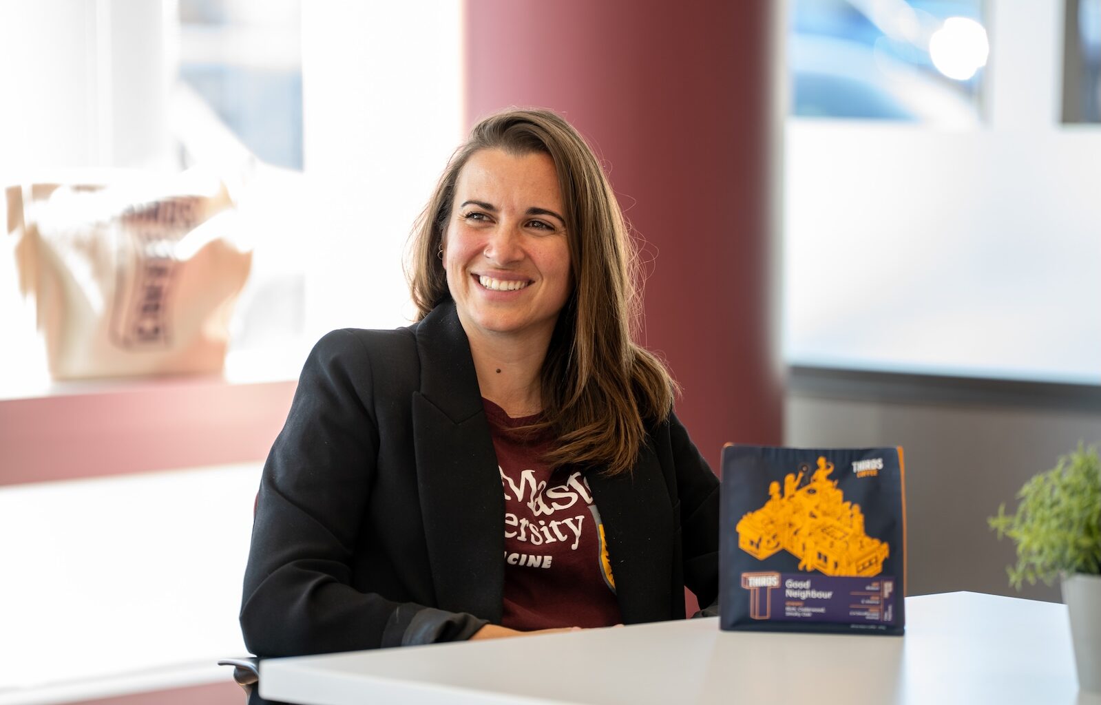 A woman in a blazer sits at a table, with a bag of coffee grounds on the table in front of her.