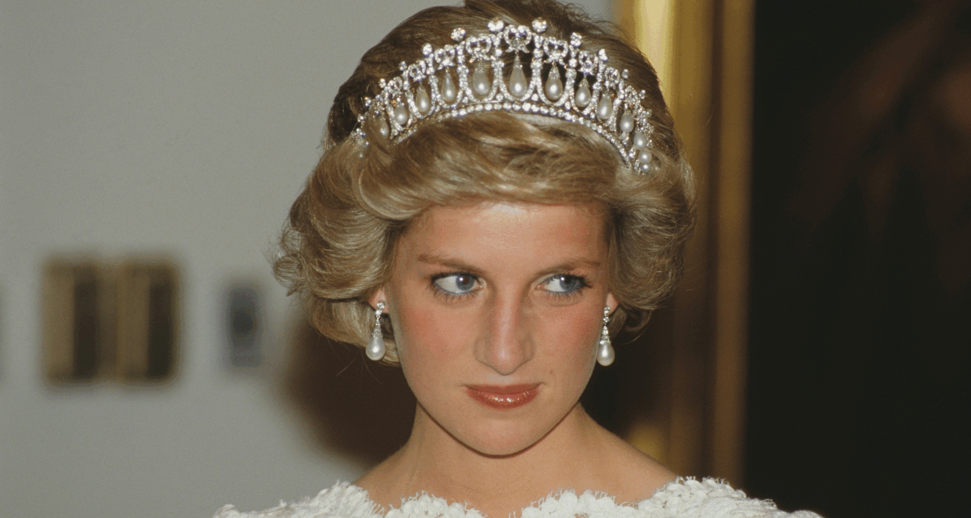 A headshot of Princess Diana wearing a tiara and pearl earrings with a white lace dress