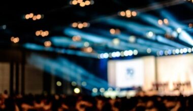 Out of focus interior of a venue, lights over an empty stage set for a panel, in front of a crowd.