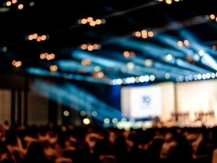 Out of focus interior of a venue, lights over an empty stage set for a panel, in front of a crowd.
