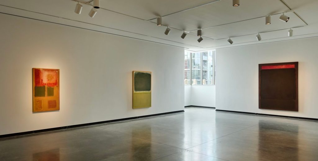 3 Rothko canvases dominated by abstract rectangular or square shapes in white-walled gallery with cement floor