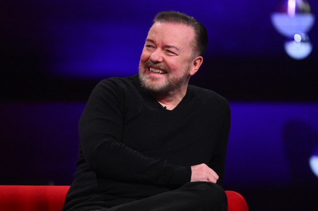 Ricky Gervais smiles during an episode of The Graham Norton Show wearing all black on the red sofa.