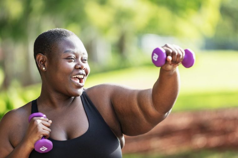 Headshot of a beautiful black woman with short hair exercising outdoors in a park on a sunny day. She is smiling, looking away, working out with dumbbell hand weights.