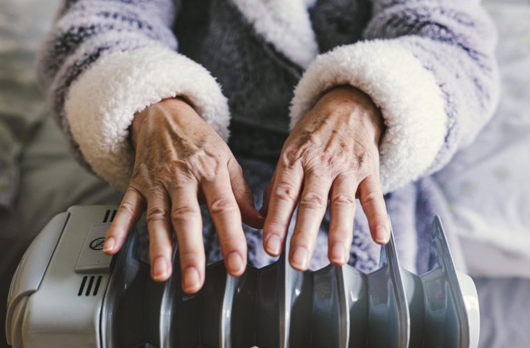A retired senior woman in her 70s sits at home inside her cold house in winter. It is so cold that she is wrapped up in warm winter clothing, and is holding her hands over an electric heater for some extra warmth and comfort. Selective focus with room for copy space.