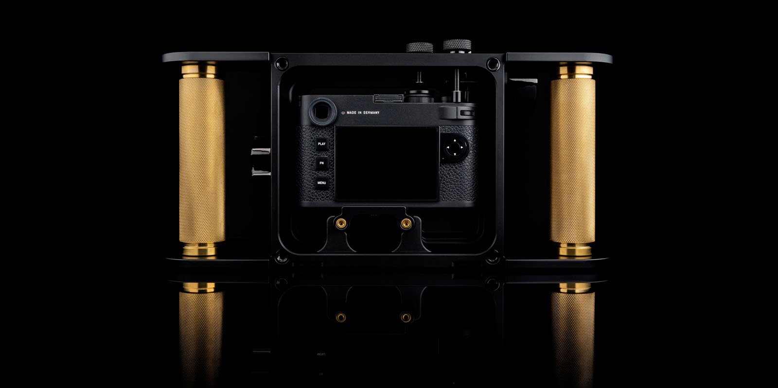 A professional camera rig with gold cylindrical grips on each side, featuring a mounted black camera centered within a rectangular black frame, displayed against a black background with a reflective surface.